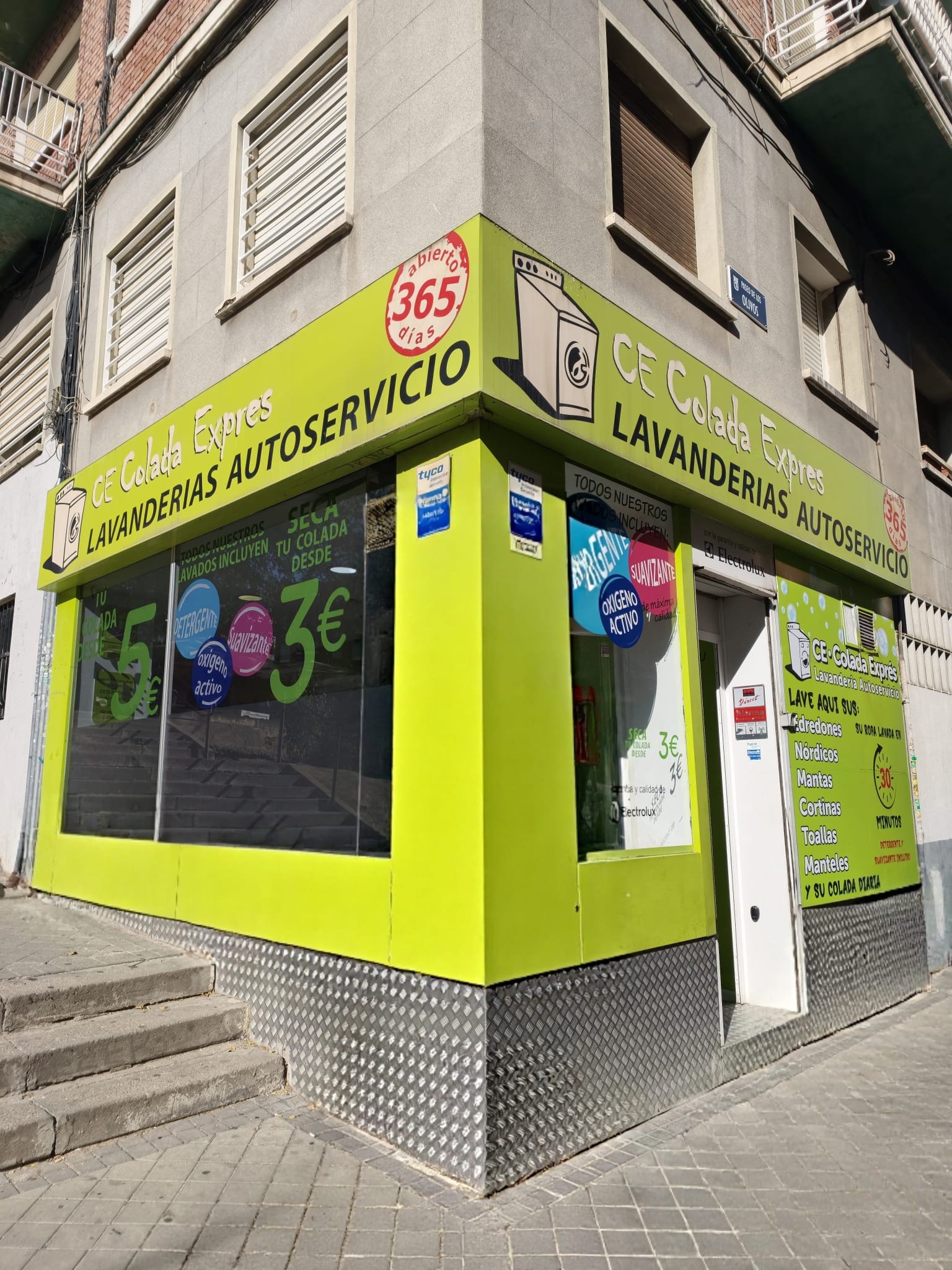 Fachada en esquina de Colada Exprés, lavandería autoservicio en el barrio de Lucero, Madrid, con el rótulo verde lima y abierto los 365 días